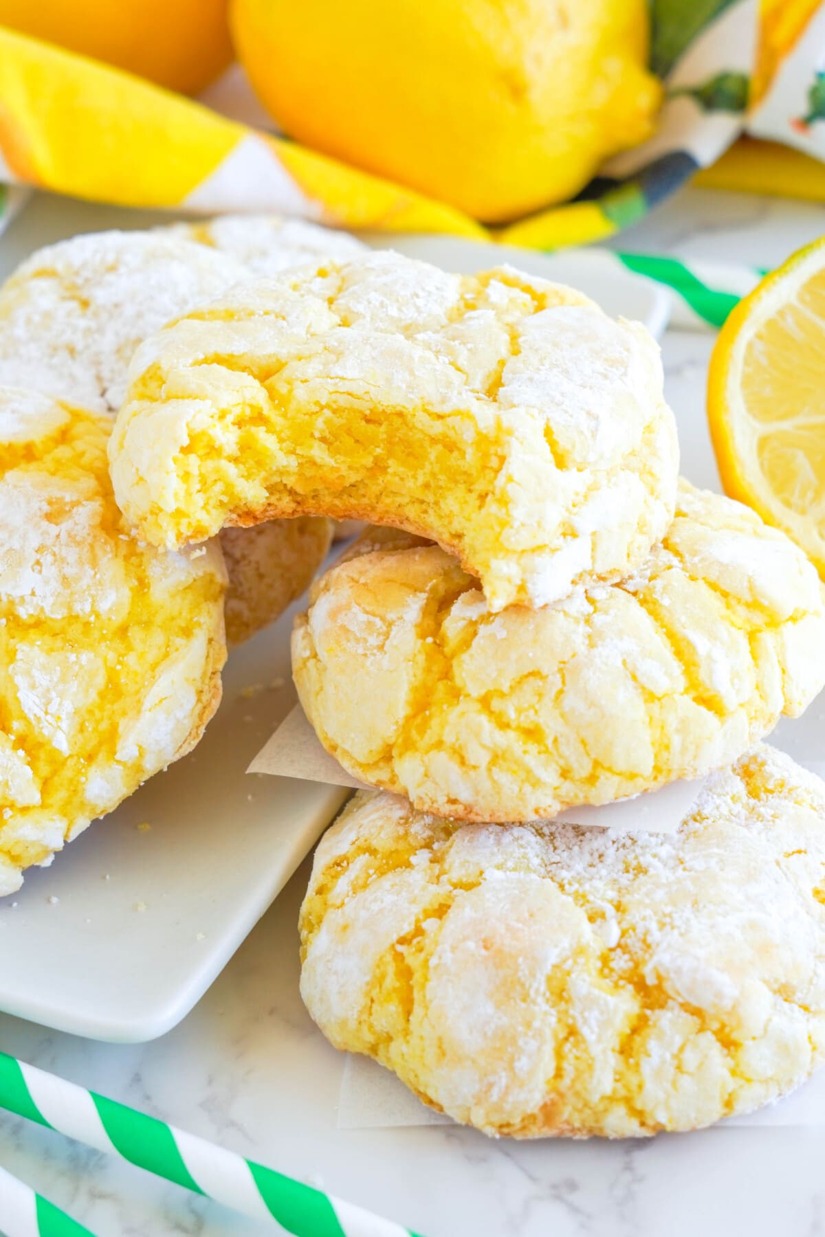 lemon crinkle cookies stacked with bite out on white plate with lemons and green striped straws