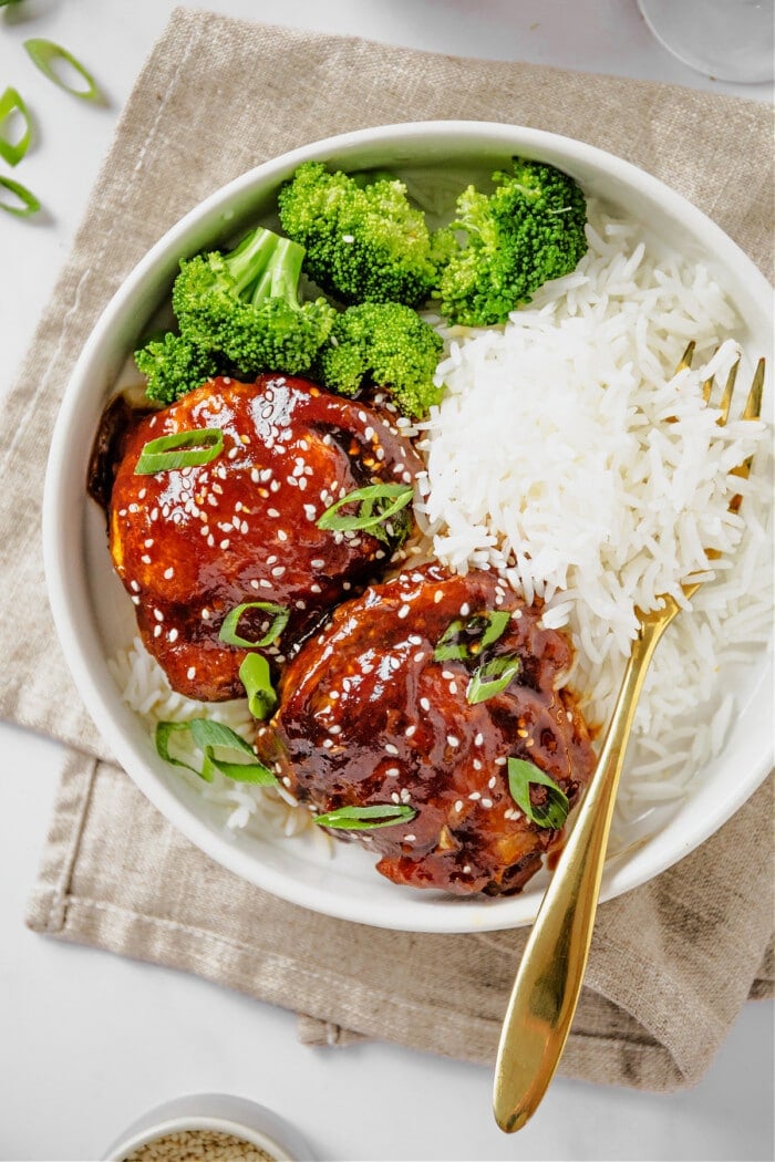 chicken thighs in white bowl with gold fork, broccoli and white rice on the side