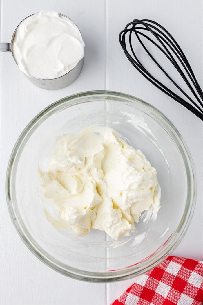 cream cheese in clear glass bowl with whisk and sour cream in measuring cup