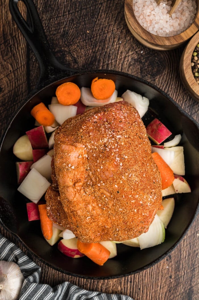 Seasoned picnic roast on top of carrots, potatoes, and onions in a cast iron skillet