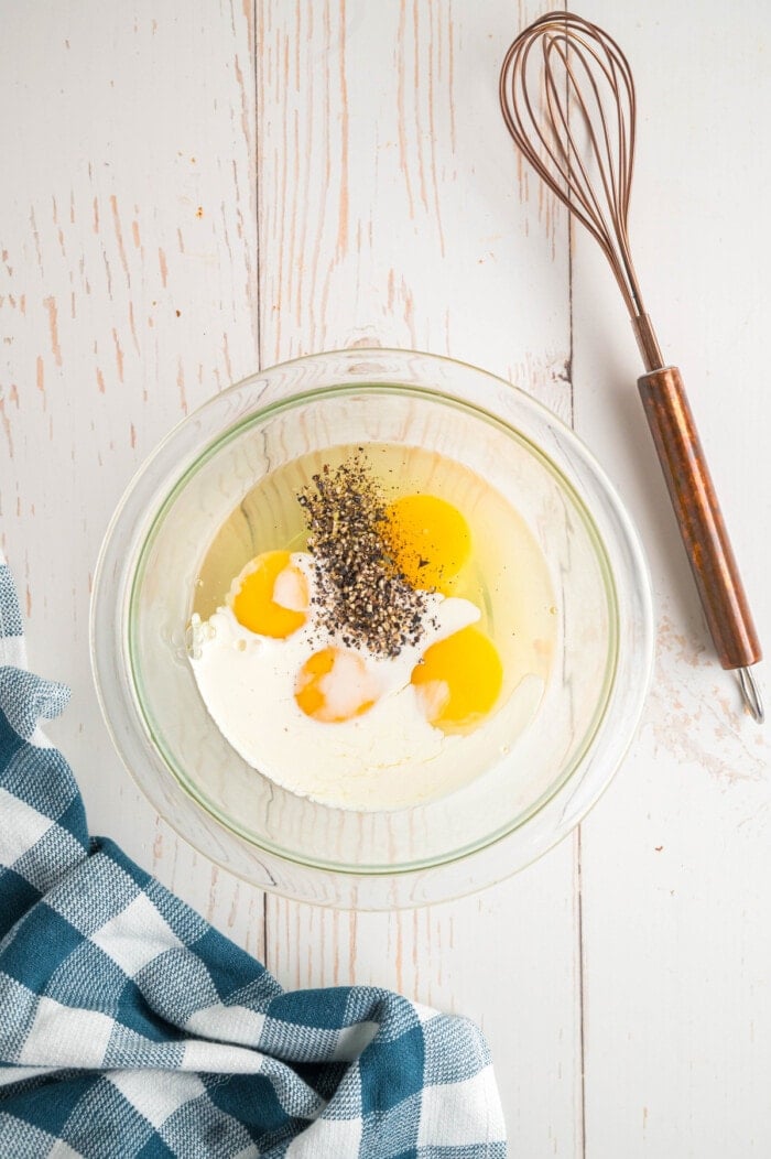 Eggs with milk and seasonings in a bowl with a whisk to the side.