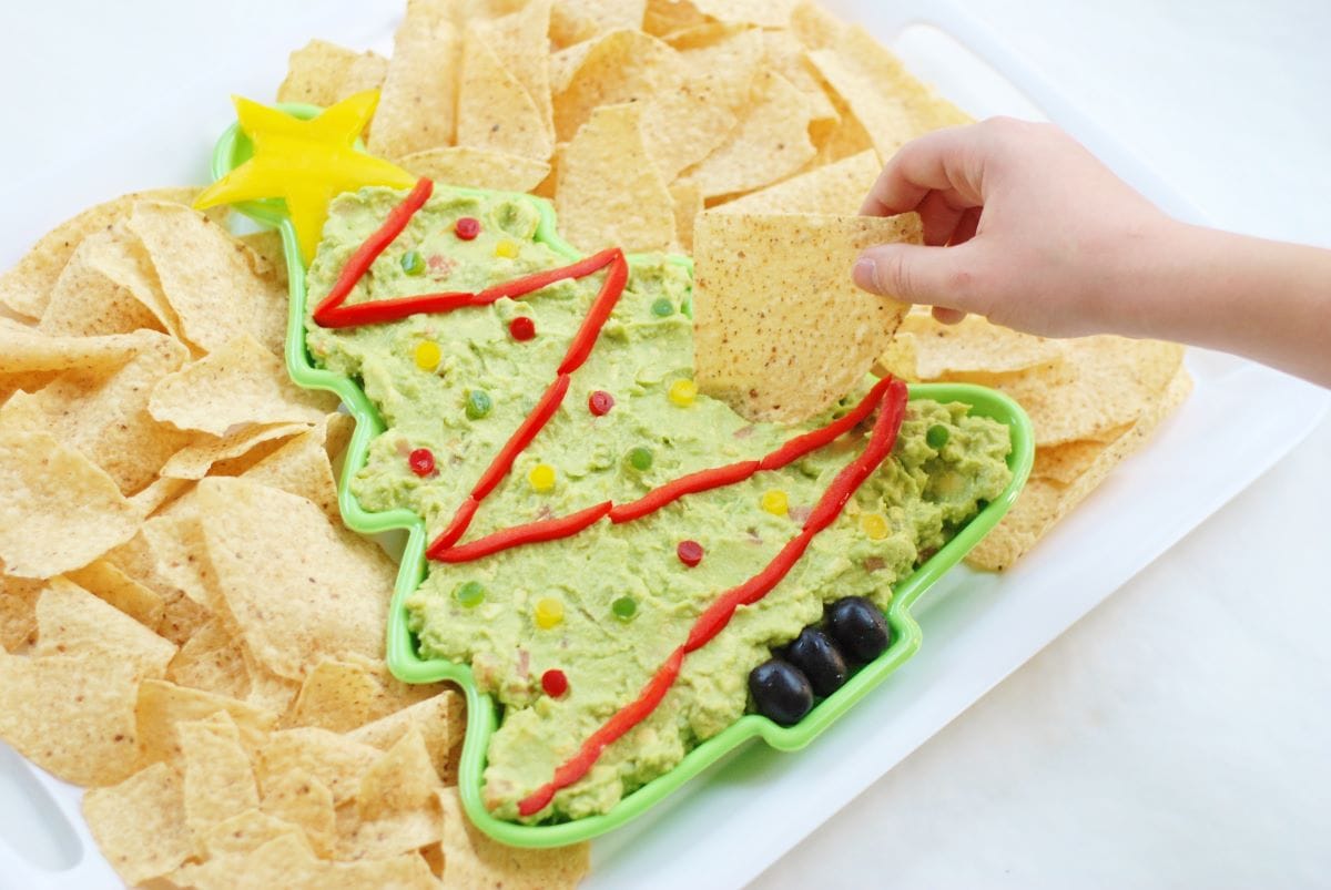 hand with a chip dipping into a Guacamole Christmas Tree surrounded by tortilla chips