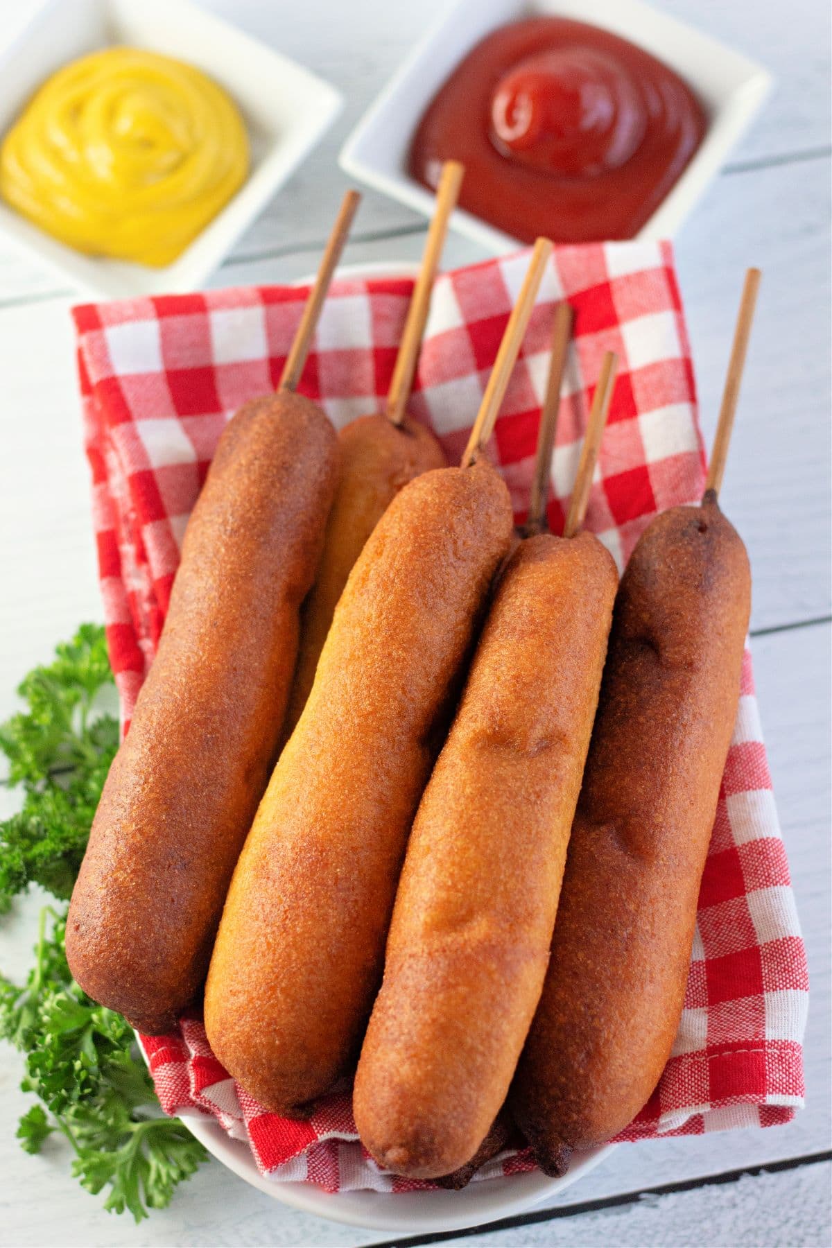six corn dogs on a red and white checkerboard cloth with ketchup and mustard in dishes