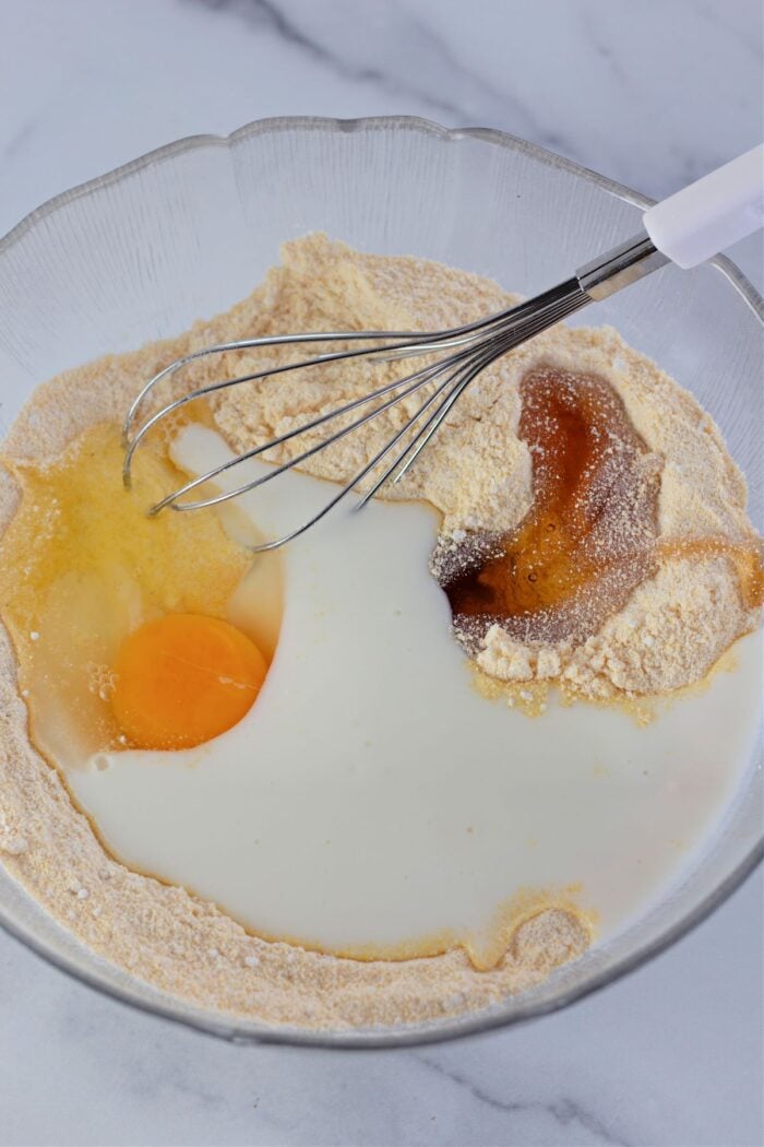whisking together cornmeal, flour, eggs, buttermilk, and honey in a clear bowl.