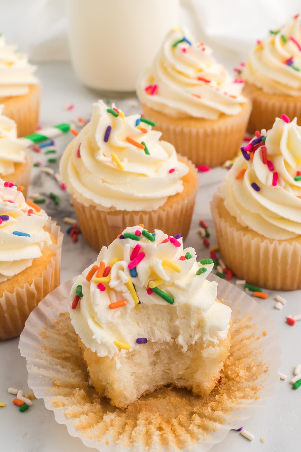 Overhead view of vanilla cupcakes topped with buttercream frosting and sprinkles, one with a bite missing.