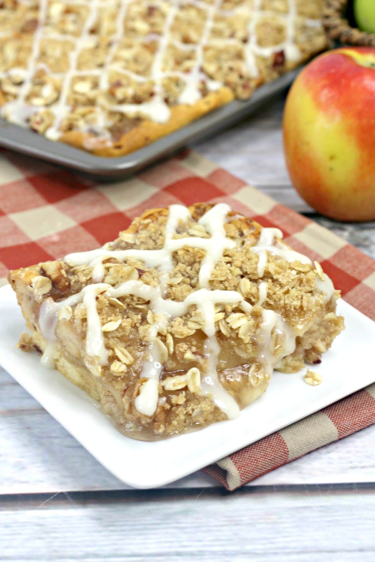A slice of apple streusel slab pie on a square plate with the rest of the pie in the background.
