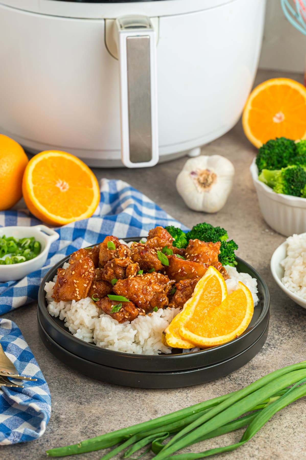 Orange chicken garnished with green onions on top of white rice on a dark plate sitting in front of a white air fryer.