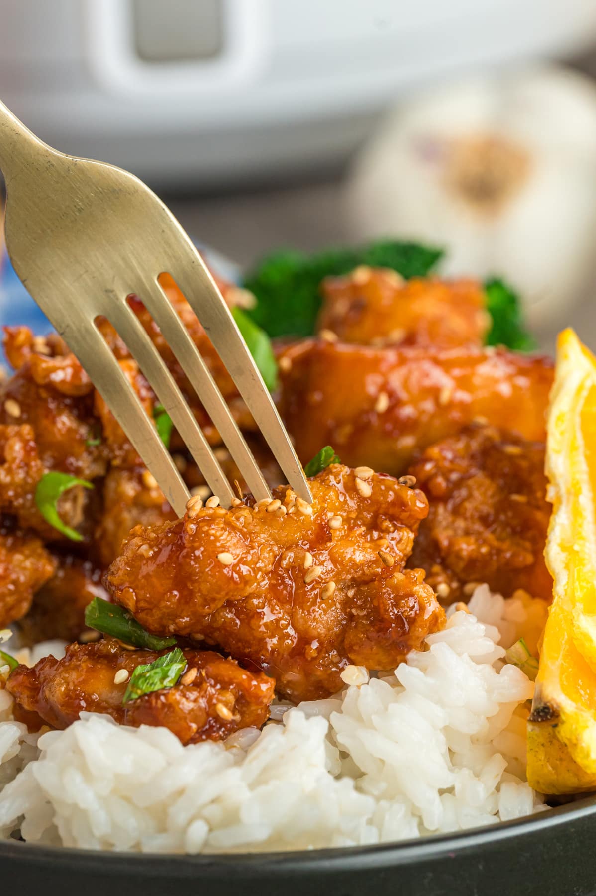 A fork picking up a piece of Orange chicken garnished with green onions on top of white rice.