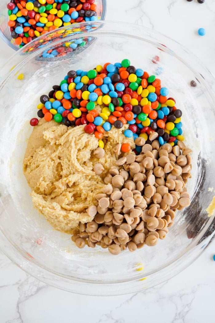 Peanut butter chips and MM's added to cookie batter in a clear bowl.