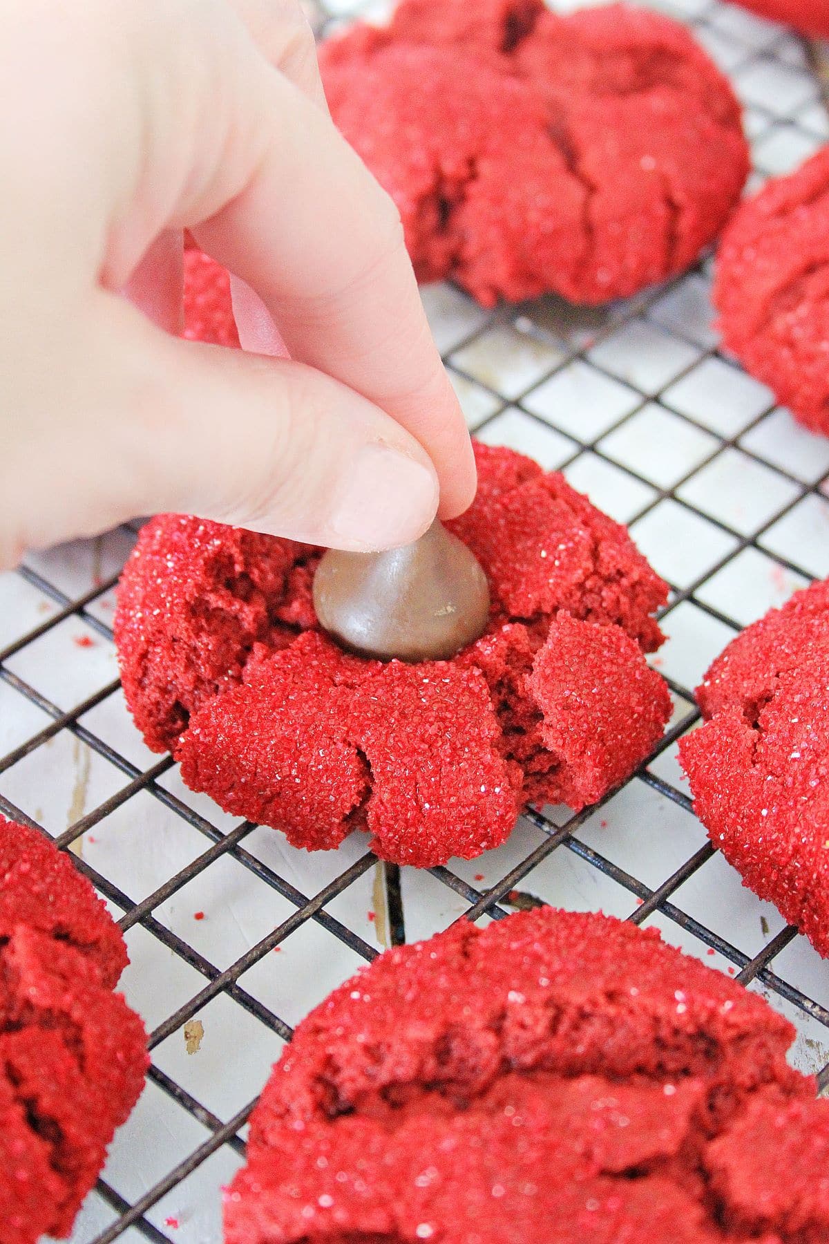 A hand placing a chocolate kiss in the center of a red velvet peanut butter cookie.