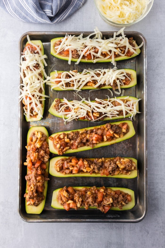 Overhead view of stuffed zucchini boats on a baking sheet, half of them topped with cheese