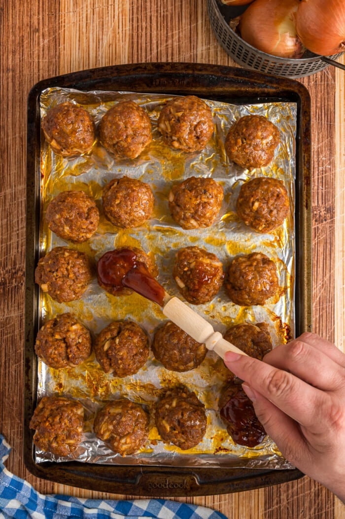 BBQ sauce being brushed over baked meatballs on a baking sheet