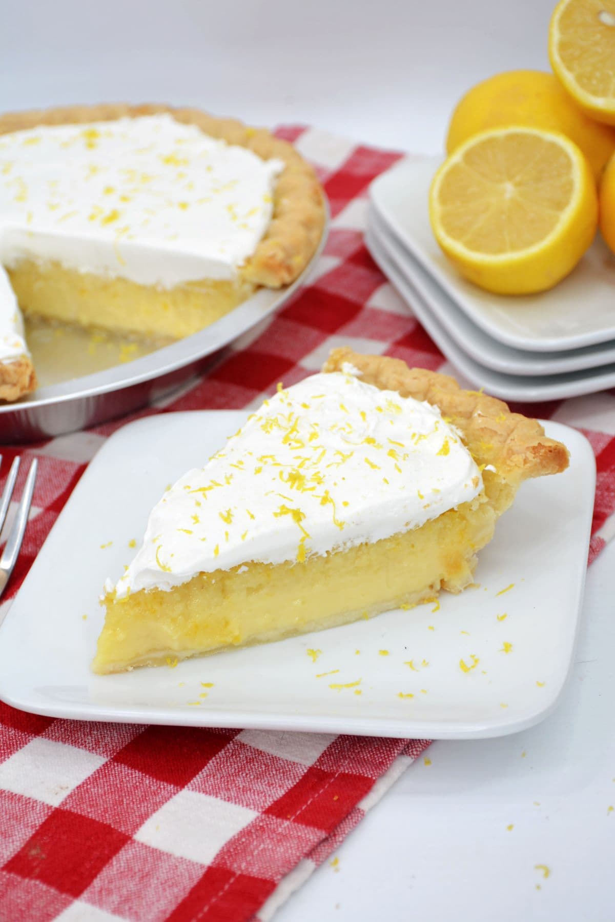 A slice of Blender Lemon Pie on a white serving plate with the full and pie and a plate of lemons in the background.