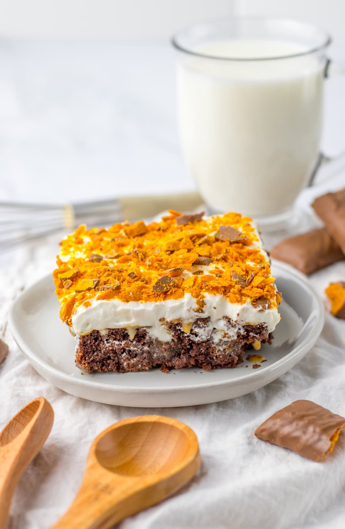 With this Butterfinger Poke Cake, you get a rich chocolate cake filled with creamy peanut butter and caramel. Butterfinger Poke Cake on white plate with glass of milk