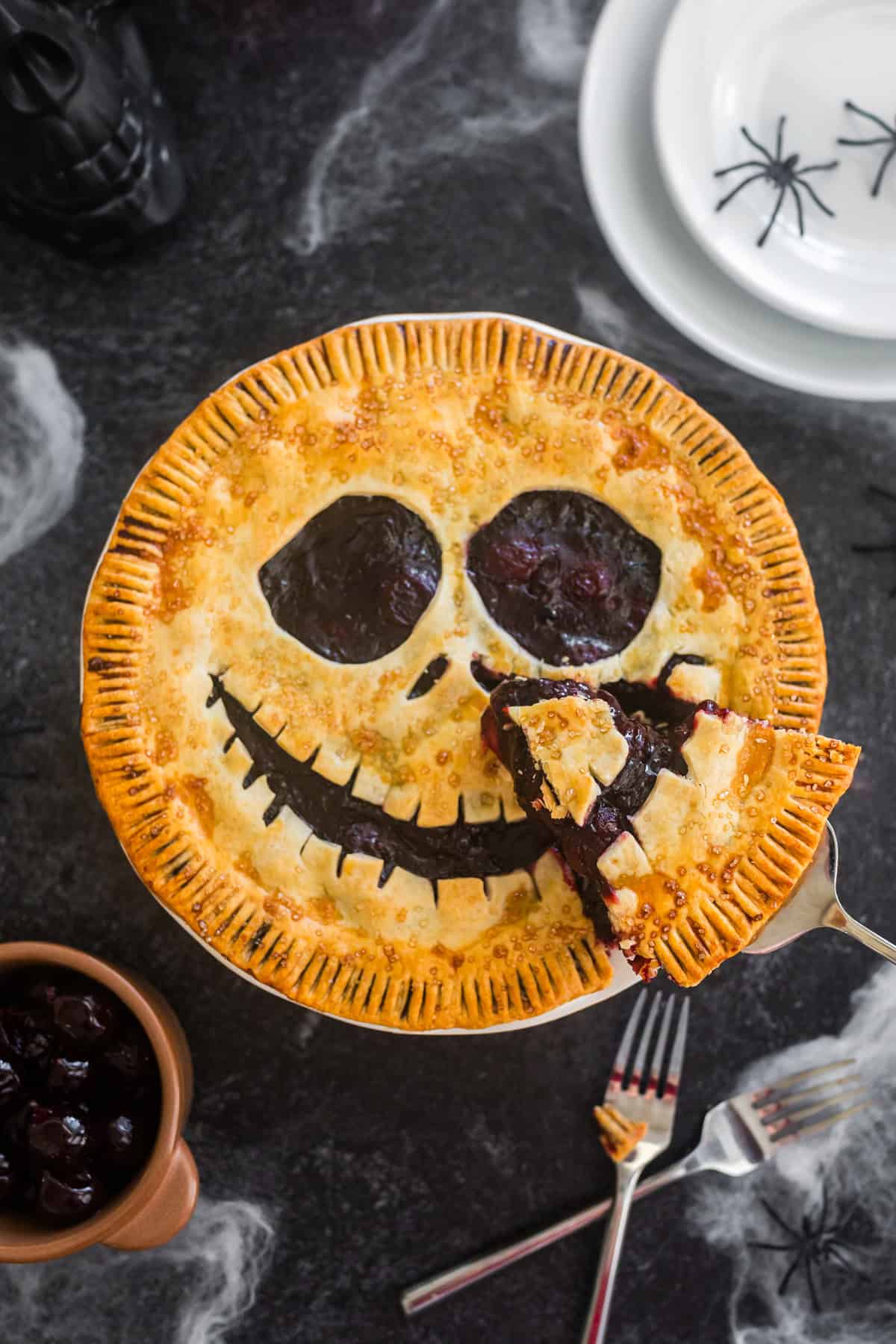 This spooky Halloween Pie is a festive take on a classic cherry pie. The deep red color of the cherries and the Jack Skellington pie crust makes it perfect for the fall holiday. Overhead look at a spooky face cherry pie while serving a slice.
