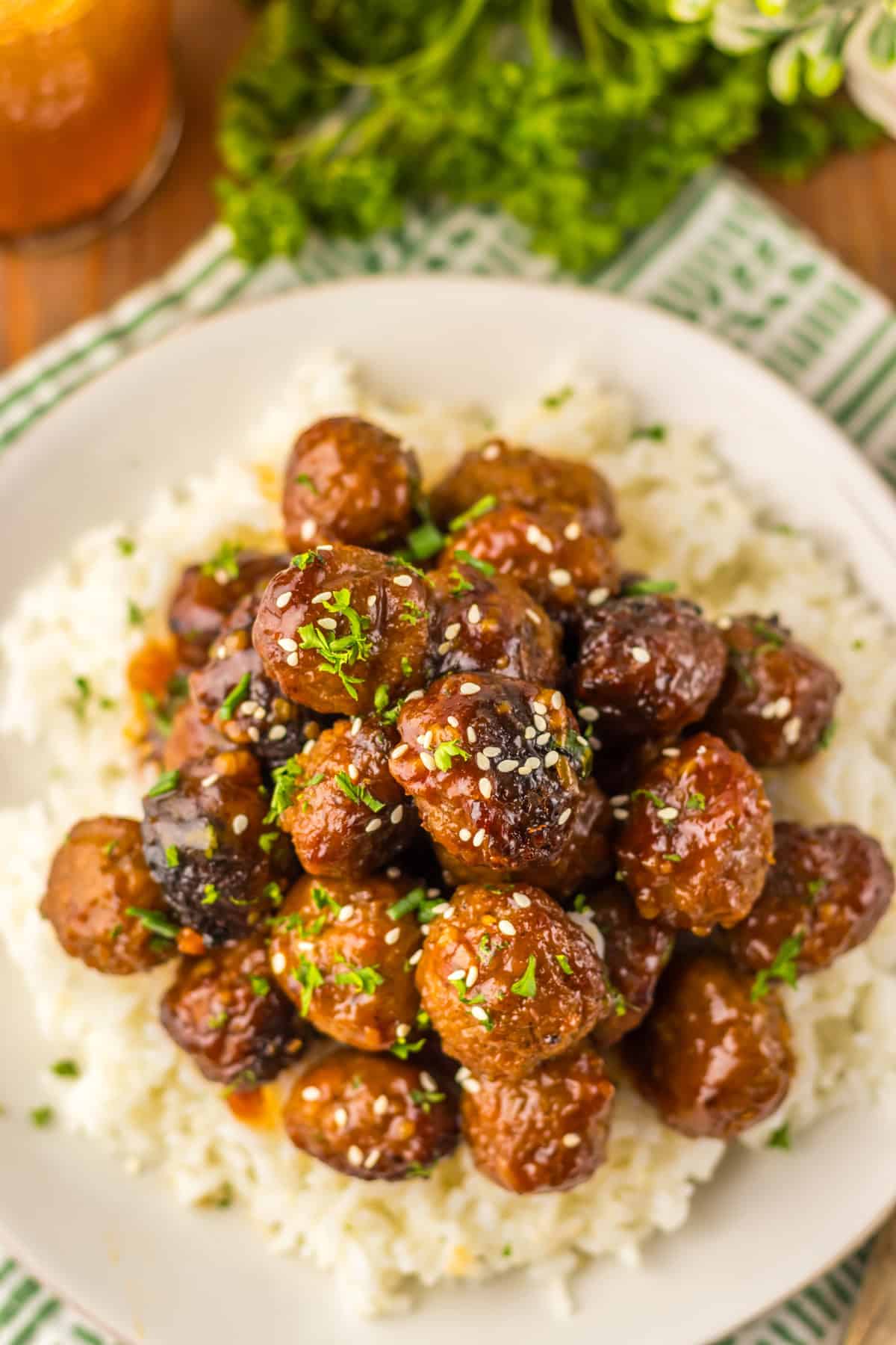 A close-up of honey garlic meatballs piled high on a plate of white rice, topped with sesame seeds and chopped parsley. The vibrant green of the parsley contrasts with the rich brown glaze of the meatballs.