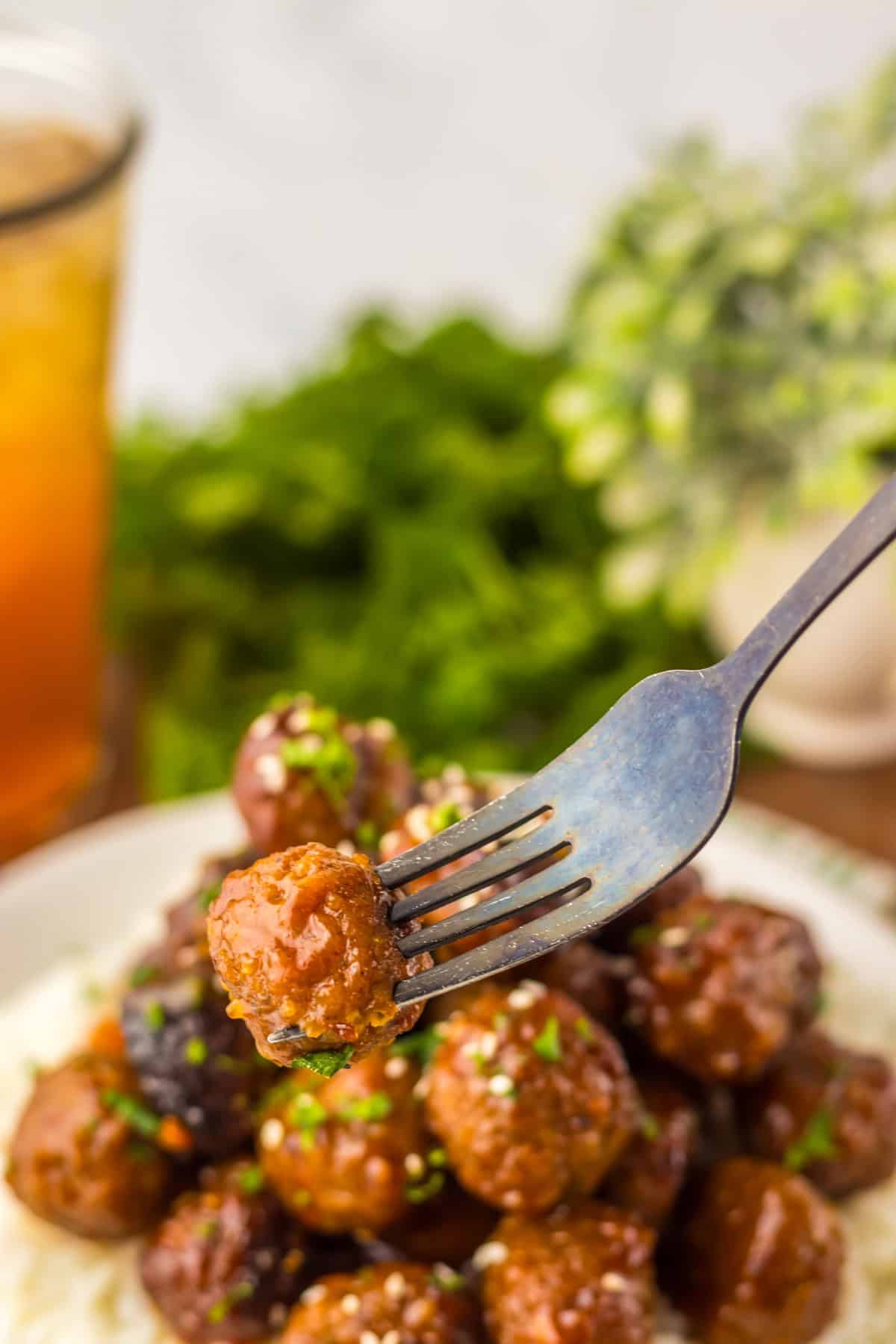 A single honey garlic meatball is held up by a fork above the plate, showcasing the shiny glaze and the bits of chopped parsley. The blurred background includes a glass of iced tea and greenery.