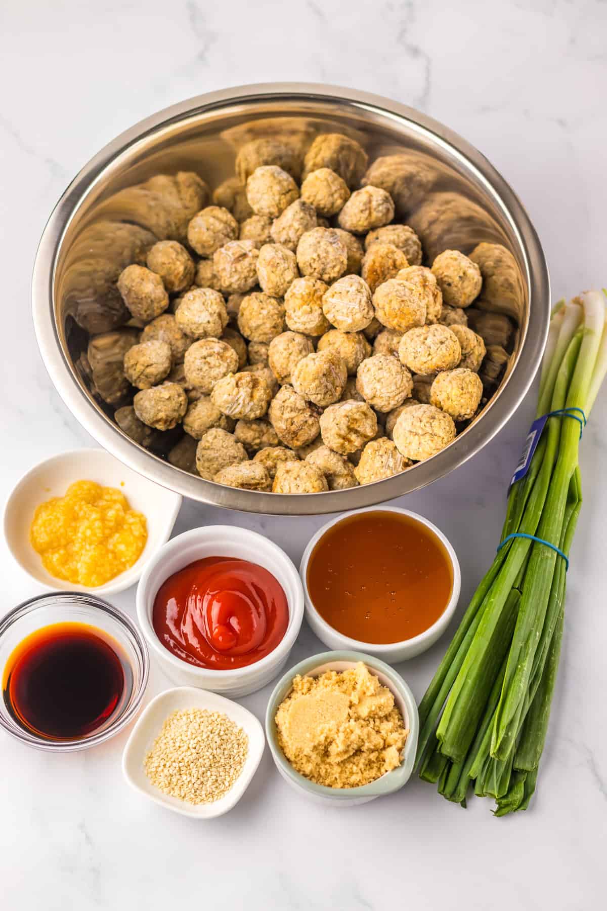 A top-down view of the ingredients for honey garlic meatballs, including a large bowl of uncooked meatballs and smaller bowls of soy sauce, garlic, ketchup, honey, brown sugar, and sesame seeds. Fresh green onions are placed alongside the bowls on a white marble surface.