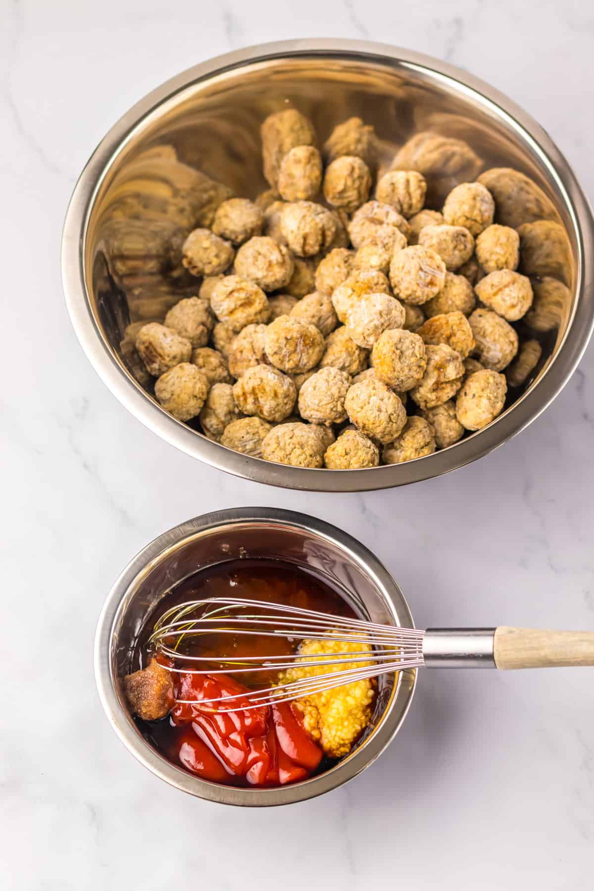 A large metal bowl filled with uncooked meatballs, alongside a smaller bowl where a whisk is mixing ketchup, honey, garlic, and other sauce ingredients. The scene is set on a white marble countertop.
