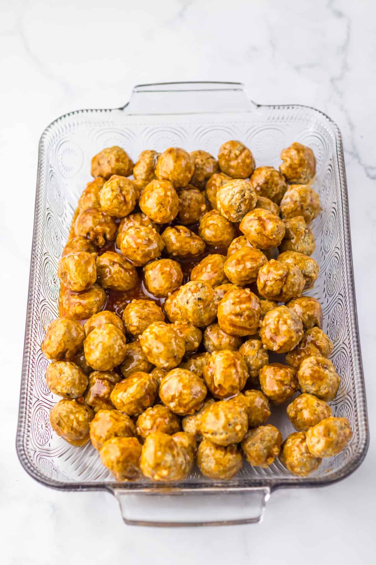 glass baking dish filled with uncooked meatballs coated in a honey garlic sauce, ready to be baked. The dish is on a white marble surface.