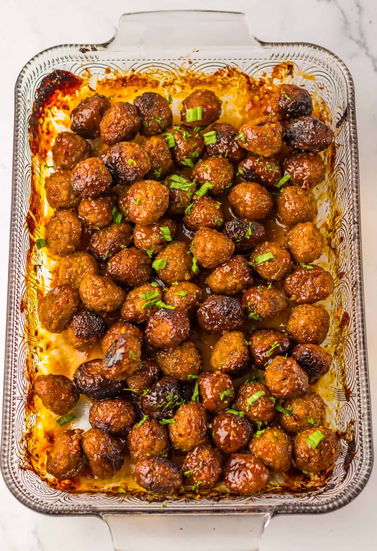 A glass baking dish filled with baked honey garlic meatballs, garnished with chopped green onions and parsley. The edges of the dish show a bit of caramelization from the baked sauce.