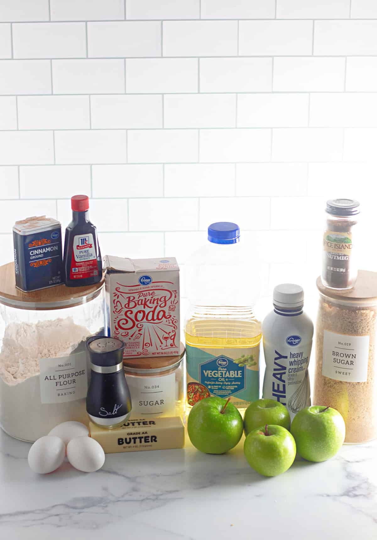 A collection of ingredients for the Apple Dapple Cake, including flour, butter, eggs, apples, vegetable oil, heavy cream, sugar, and spices, arranged on a kitchen counter.