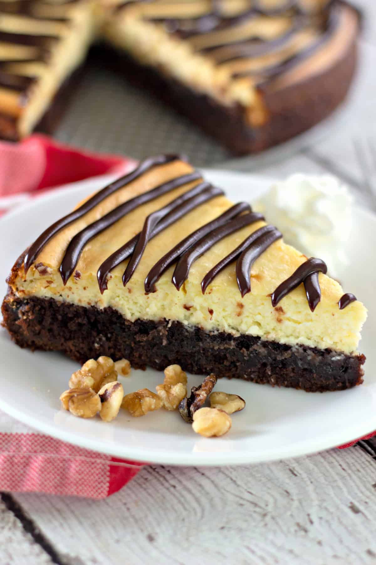 Indulge in pure dessert heaven with this homemade Brownie Bottom Cheesecake! Imagine a rich, fudgy brownie base topped with a smooth, creamy cheesecake filling—it’s the perfect combo of two classic treats in one bite. This show-stopping dessert will quickly become a family favorite, whether you're treating the kids after a long day or impressing guests at your next gathering. Overhead view of a slice of Brownie Bottom Cheesecake on a white plate with walnuts and whipped cream, next to the rest of the cheesecake and a red checkered napkin.