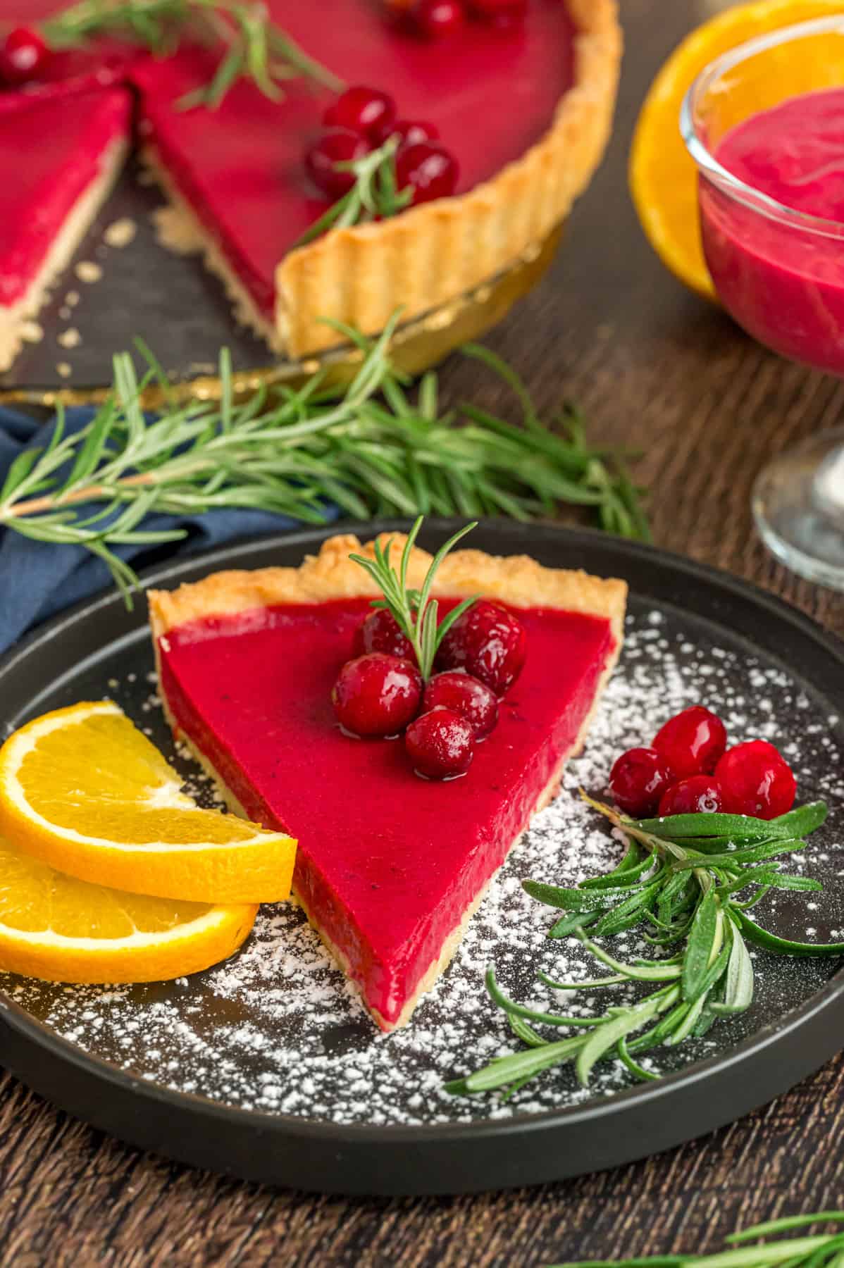 This Cranberry Curd Tart is the perfect balance of sweet and tart, made with fresh, juicy cranberries. cranberry curd tart with fresh cranberries and rosemary on top served on a gray plate with fresh oranges