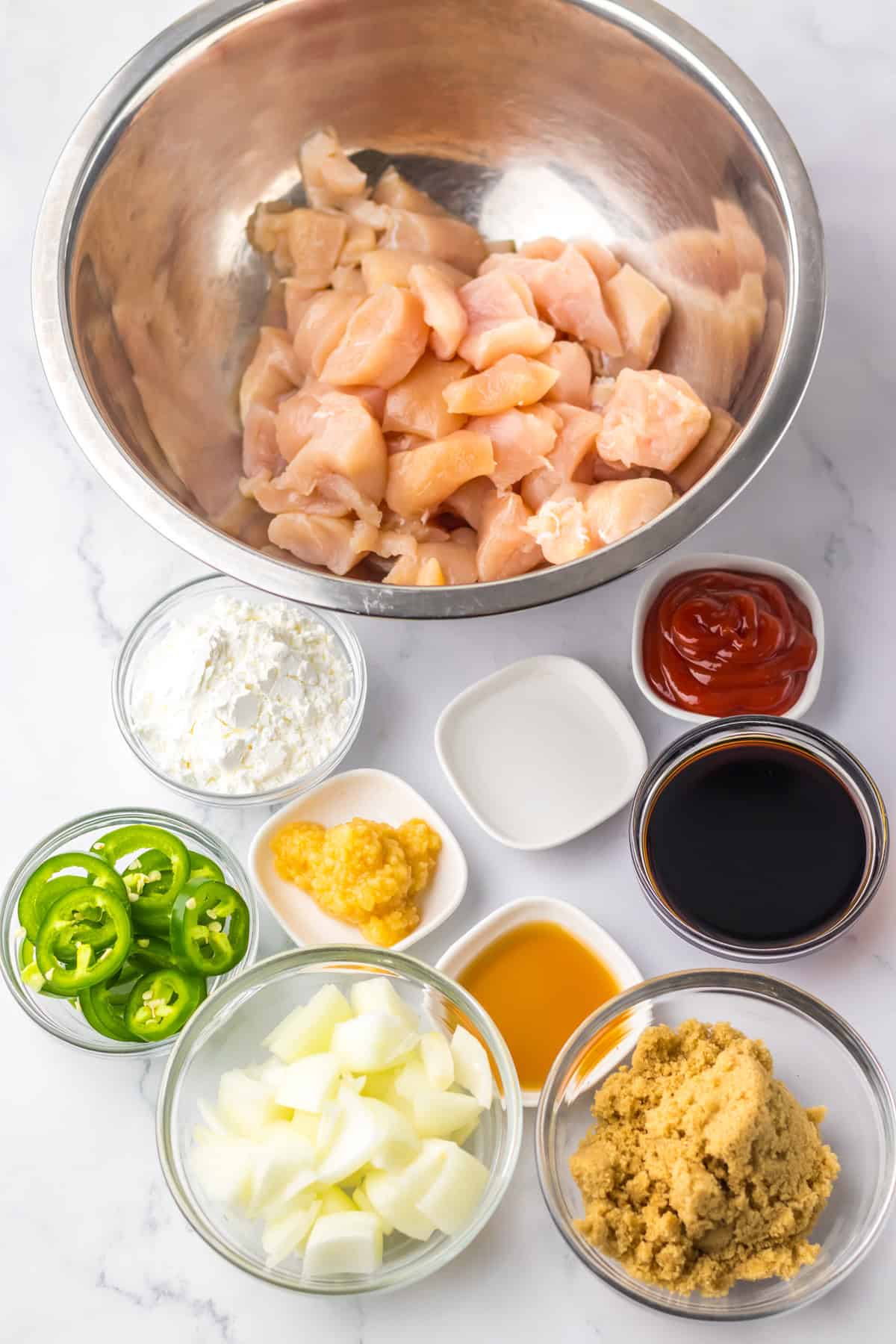 Prepped ingredients for jalapeño chicken displayed in small bowls, including raw chicken, sliced jalapeños, chopped onions, minced garlic, ketchup, soy sauce, vinegar, brown sugar, cornstarch, and oil.