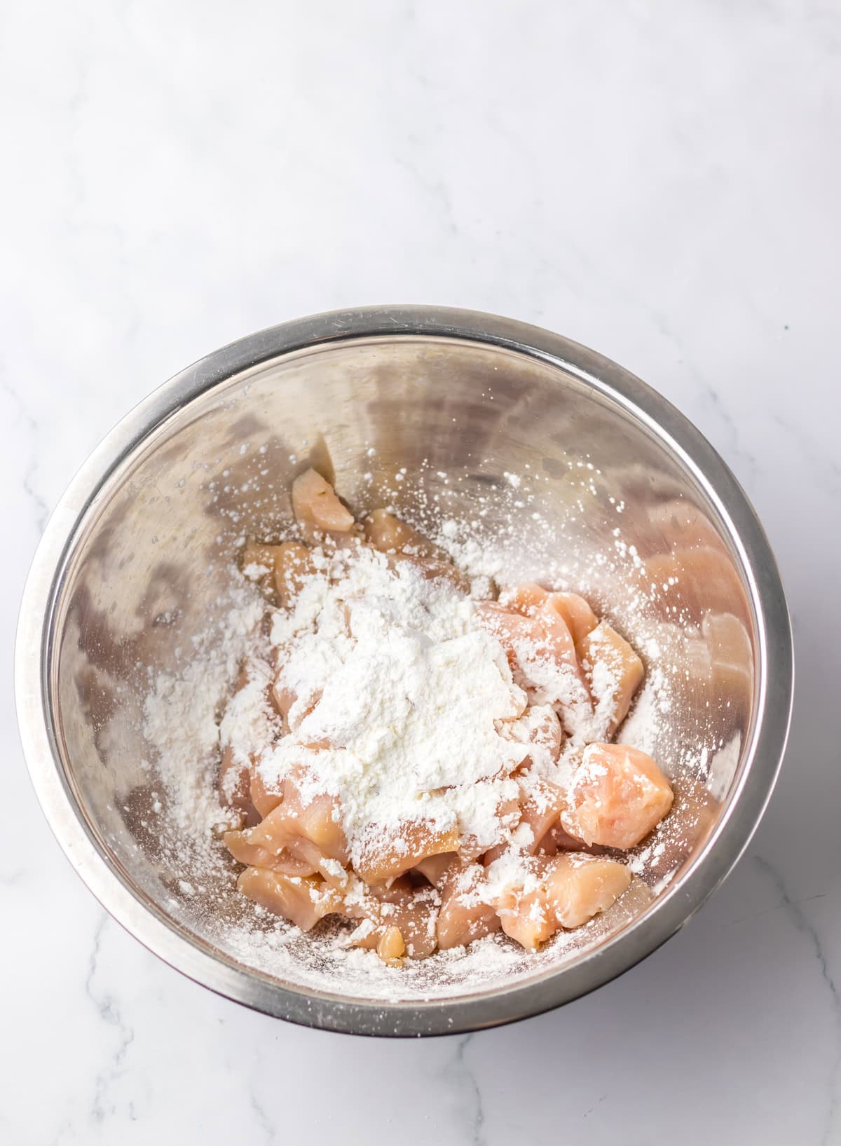 Raw chicken pieces being coated with flour or cornstarch in a large metal mixing bowl, part of the preparation for jalapeño chicken.