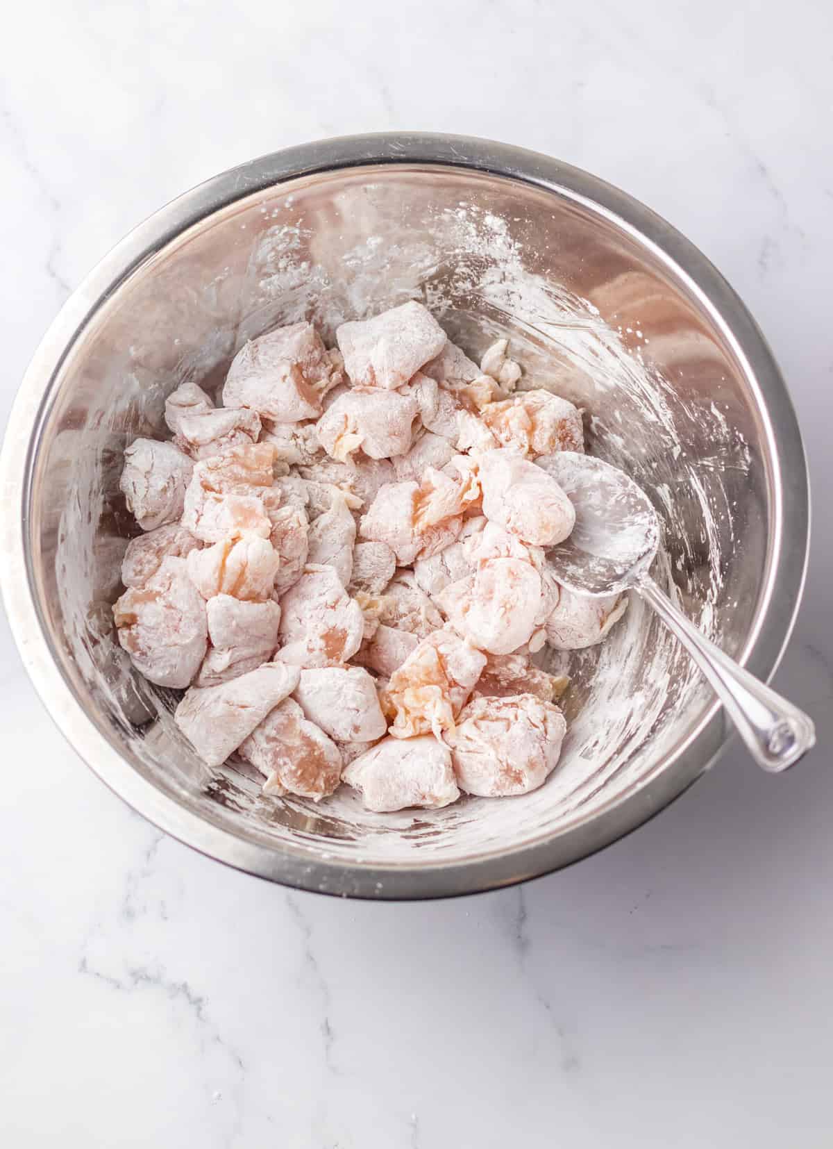 Raw chicken pieces coated in flour or cornstarch inside a large metal mixing bowl with a spoon, ready to be fried for jalapeño chicken.
