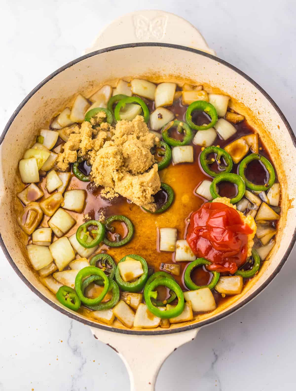 A pan with raw ingredients including onions, jalapeño slices, ketchup, and brown sugar being cooked to start the sauce for jalapeño chicken.