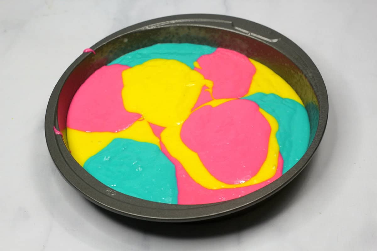 Bright tie-dye cake batter in a round cake pan, ready to be baked, showing swirls of pink, blue, and yellow.