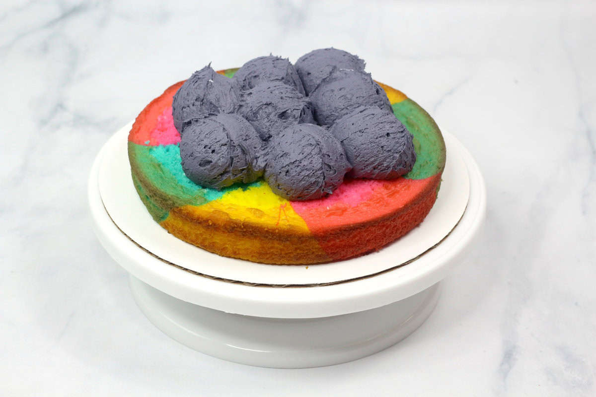Scoops of dark buttercream frosting placed on top of a baked tie-dye cake layer, ready to be spread.
