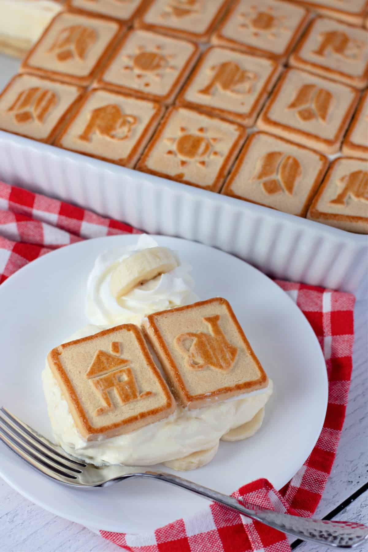 A close-up of a serving of banana pudding on a plate, featuring two Chessmen cookies on top and a dollop of whipped cream with a banana slice on the side.