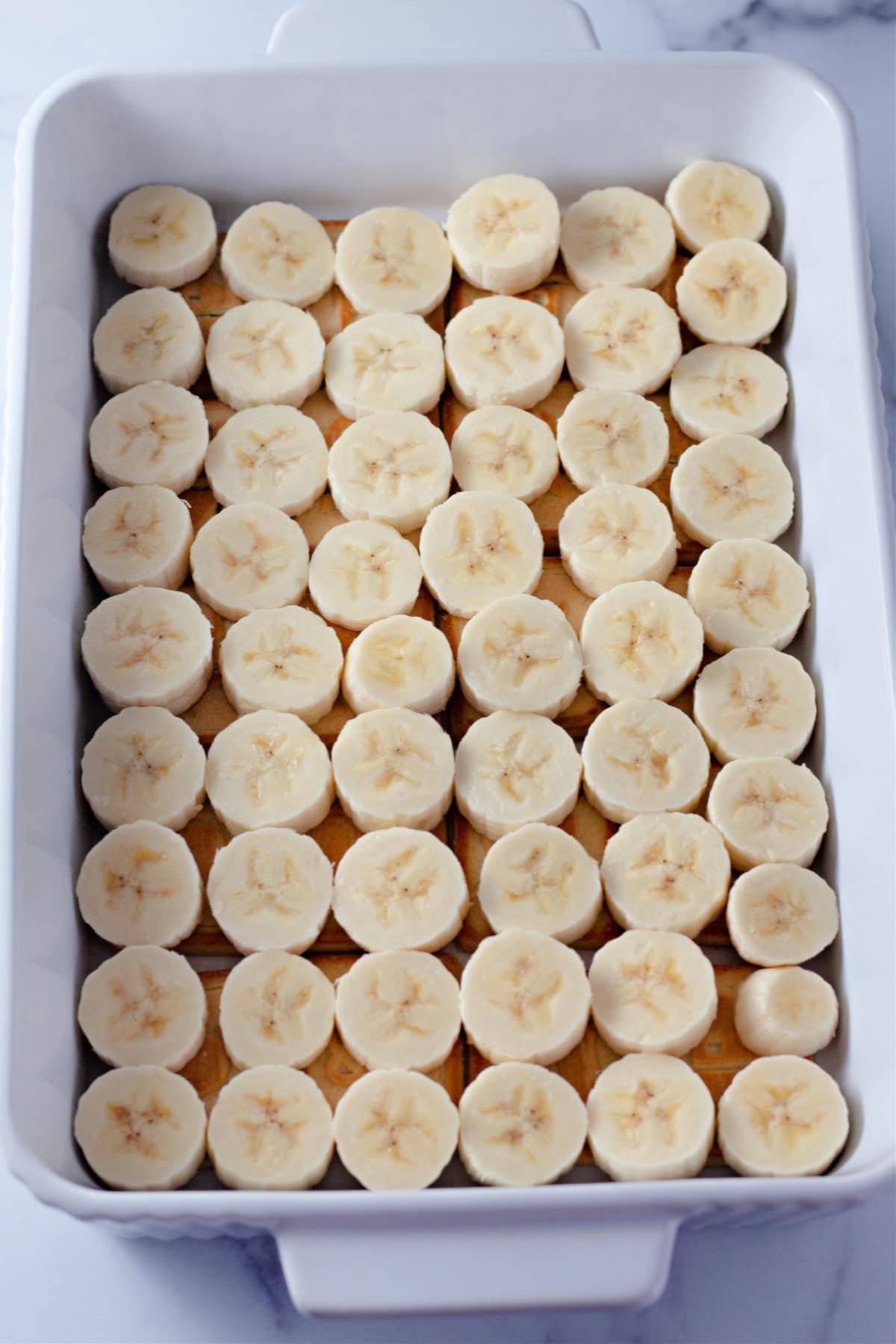 A white rectangular dish with a layer of freshly sliced banana pieces arranged over Chessmen cookies for banana pudding.