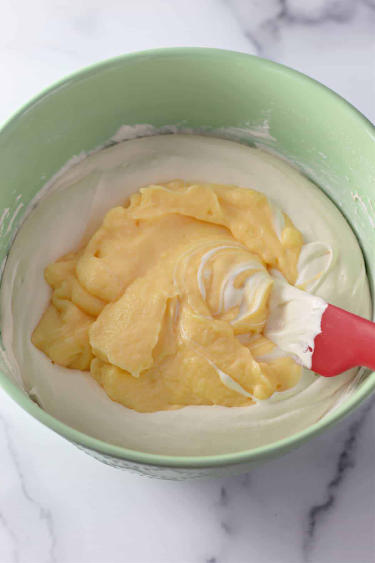 A green mixing bowl with whipped cream cheese mixture being folded into the vanilla pudding.