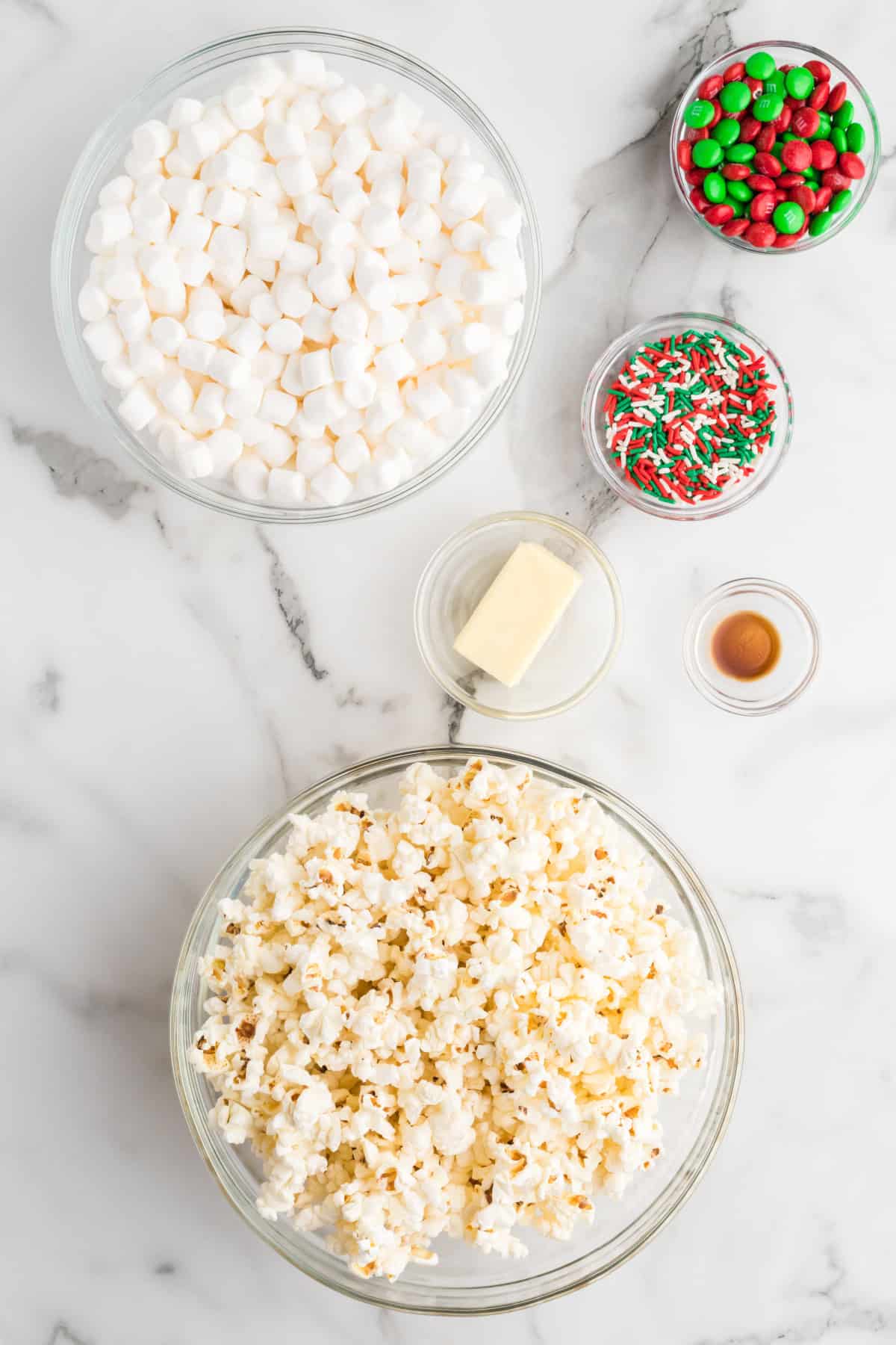 Ingredients laid out for making popcorn balls, including marshmallows, sprinkles, M&Ms, butter, and popped popcorn in clear bowls.