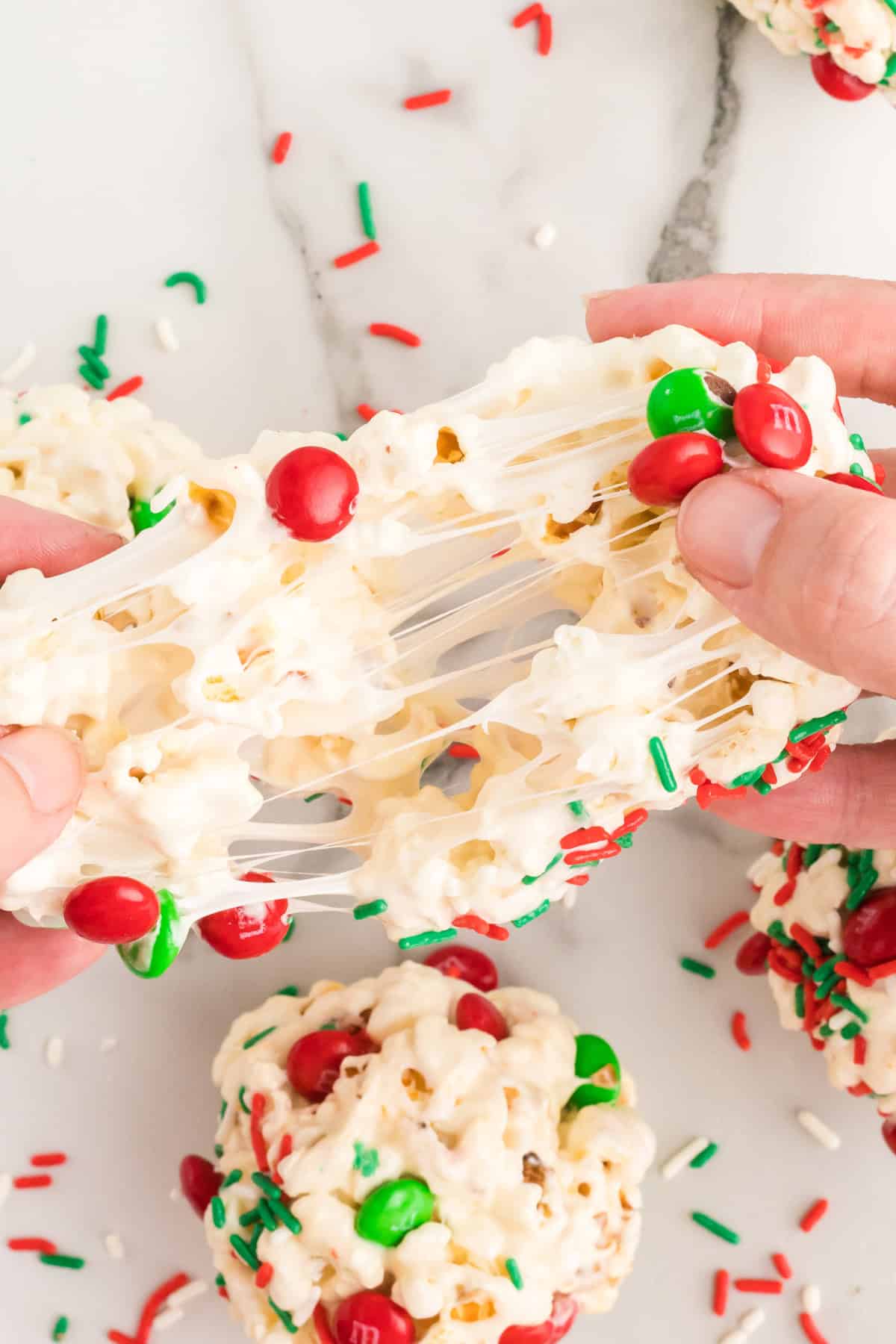 Close-up of a gooey holiday popcorn ball being stretched apart with hands, featuring colorful red and green sprinkles and M&M’s.