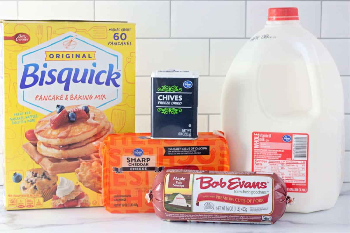 The ingredients needed to make sausage balls: Bisquick mix, shredded cheddar cheese, sausage, chives, and milk, displayed on a kitchen counter.