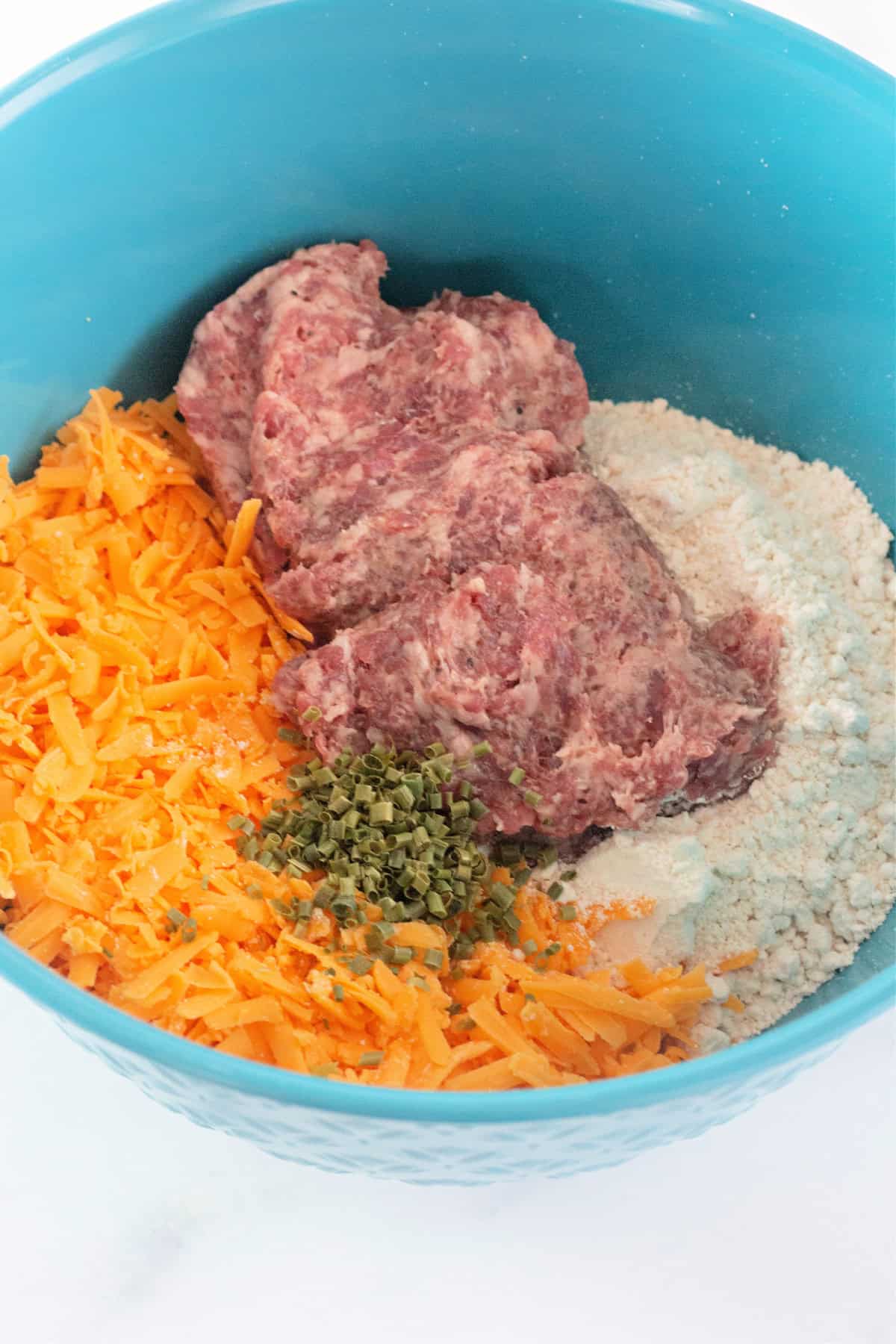 A bowl with raw ingredients for sausage balls, including ground sausage, shredded cheese, biscuit mix, and chives.