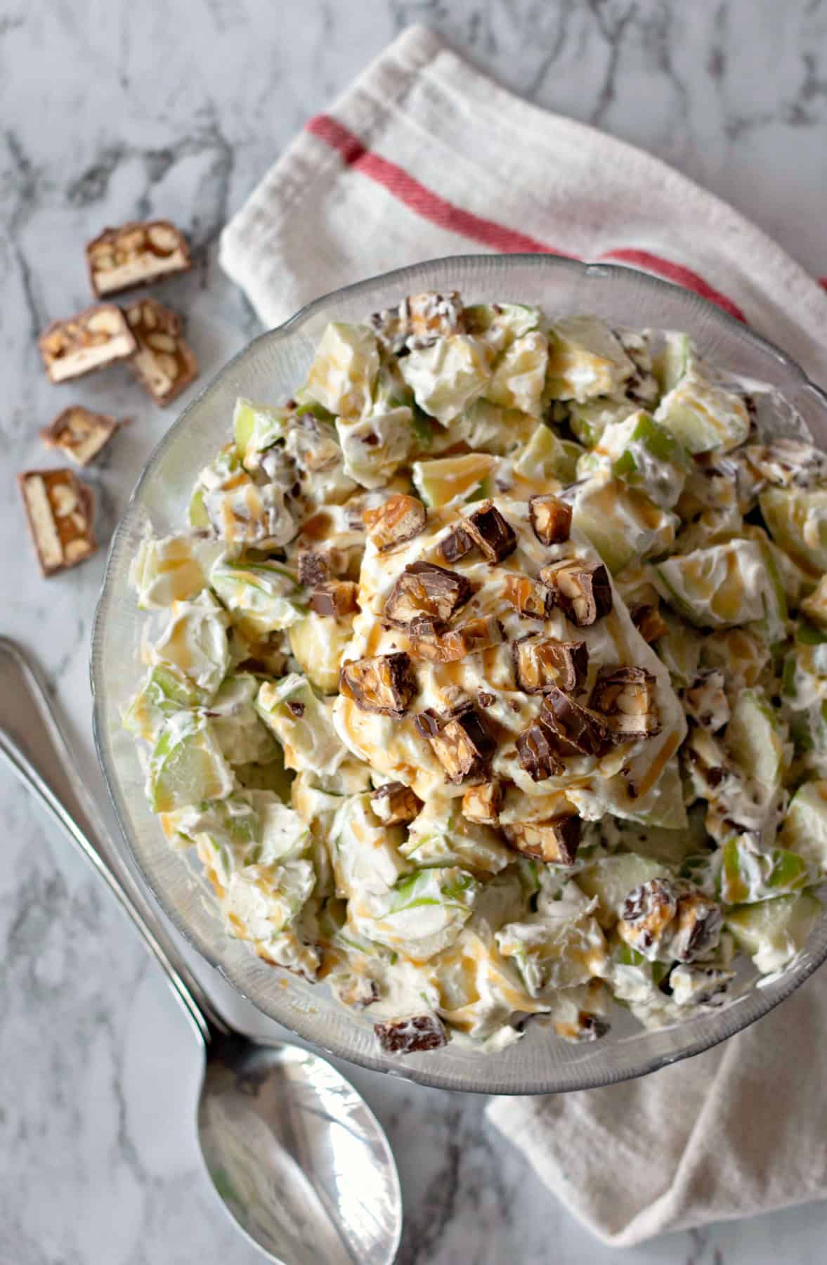 An overhead view of the complete Snickers Salad, showing the creamy apple salad topped with Snickers pieces in a glass bowl on a striped towel.
