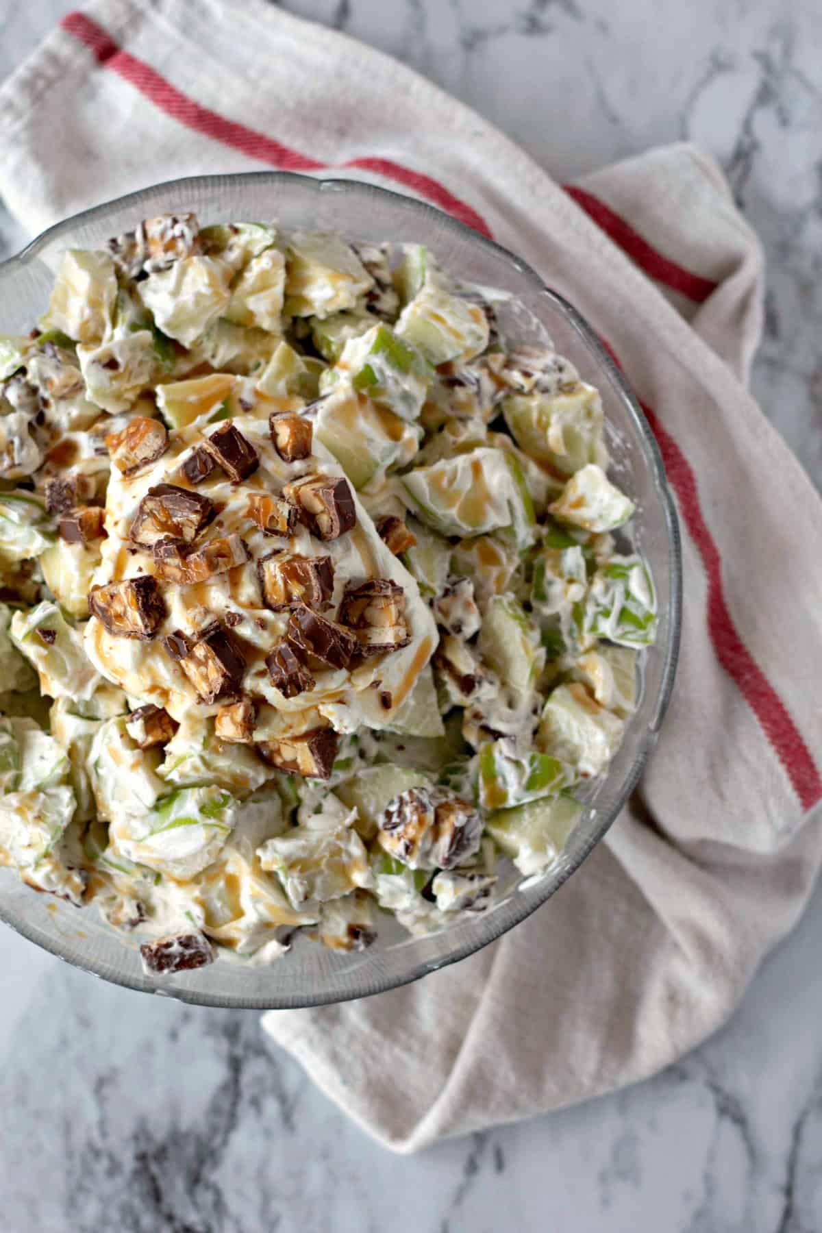 Overhead shot of Snickers Salad in a glass bowl with chunks of Snickers and apples, drizzled with caramel sauce, on a marble background with a cloth underneath.