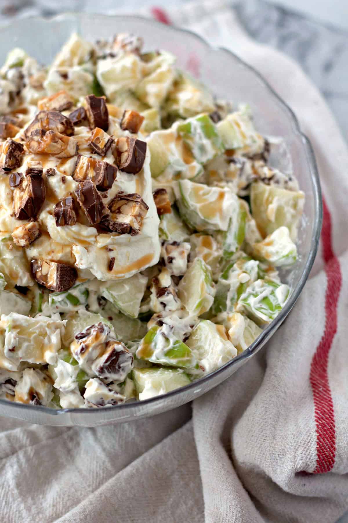 An overhead view of the complete Snickers Salad, showing the creamy apple salad topped with Snickers pieces in a glass bowl on a striped towel.