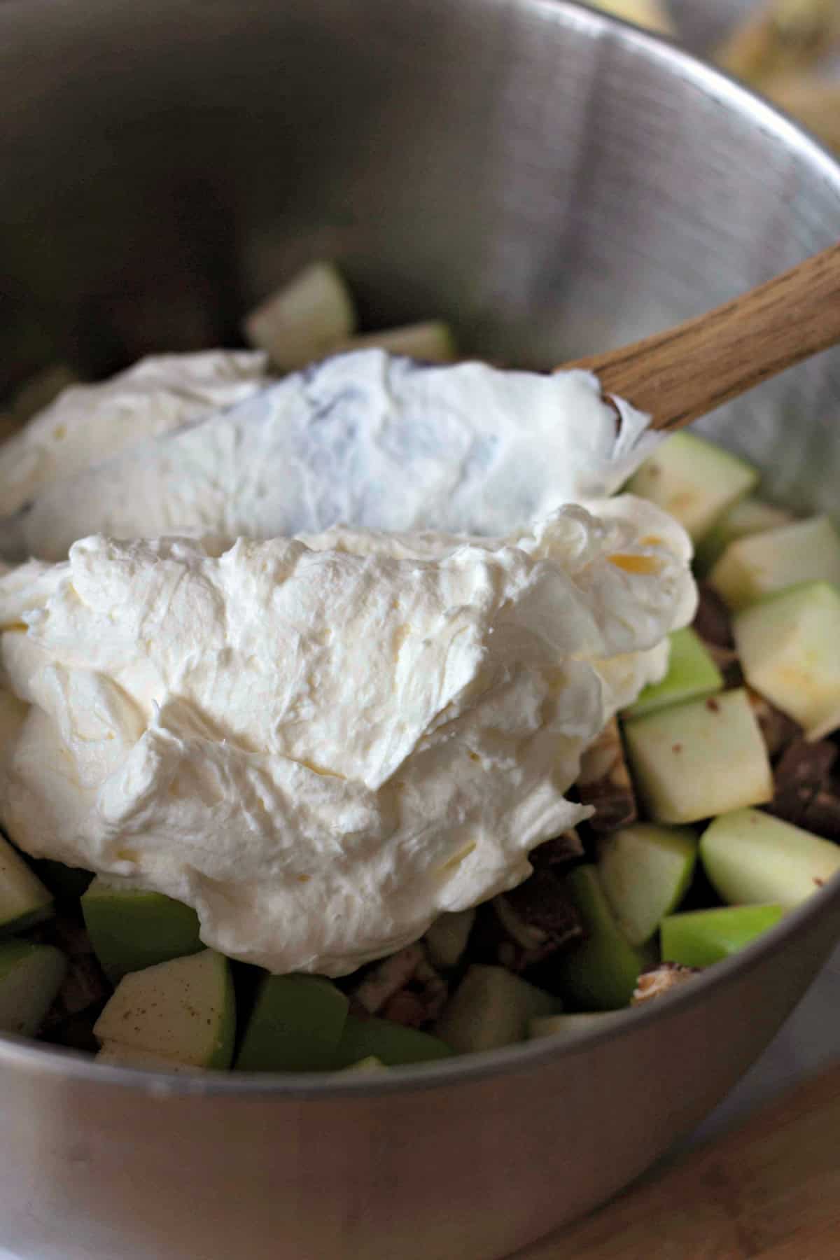 A mixing bowl with a generous scoop of whipped cream sitting on top of a mixture of chopped apples and Snickers bars, ready to be folded together.