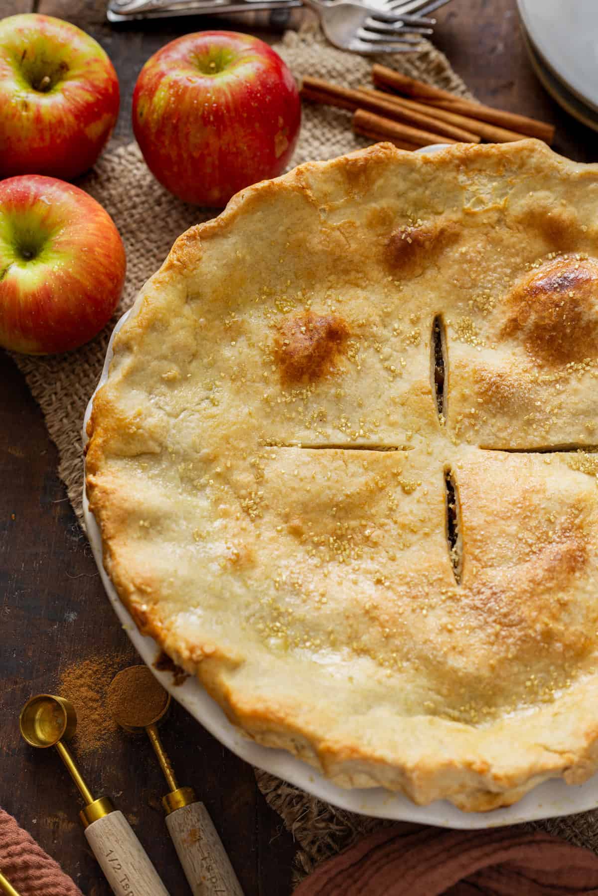 A whole, freshly baked apple pie with a golden-brown crust, garnished with sugar and cinnamon, surrounded by fresh apples and cinnamon sticks on a rustic table.