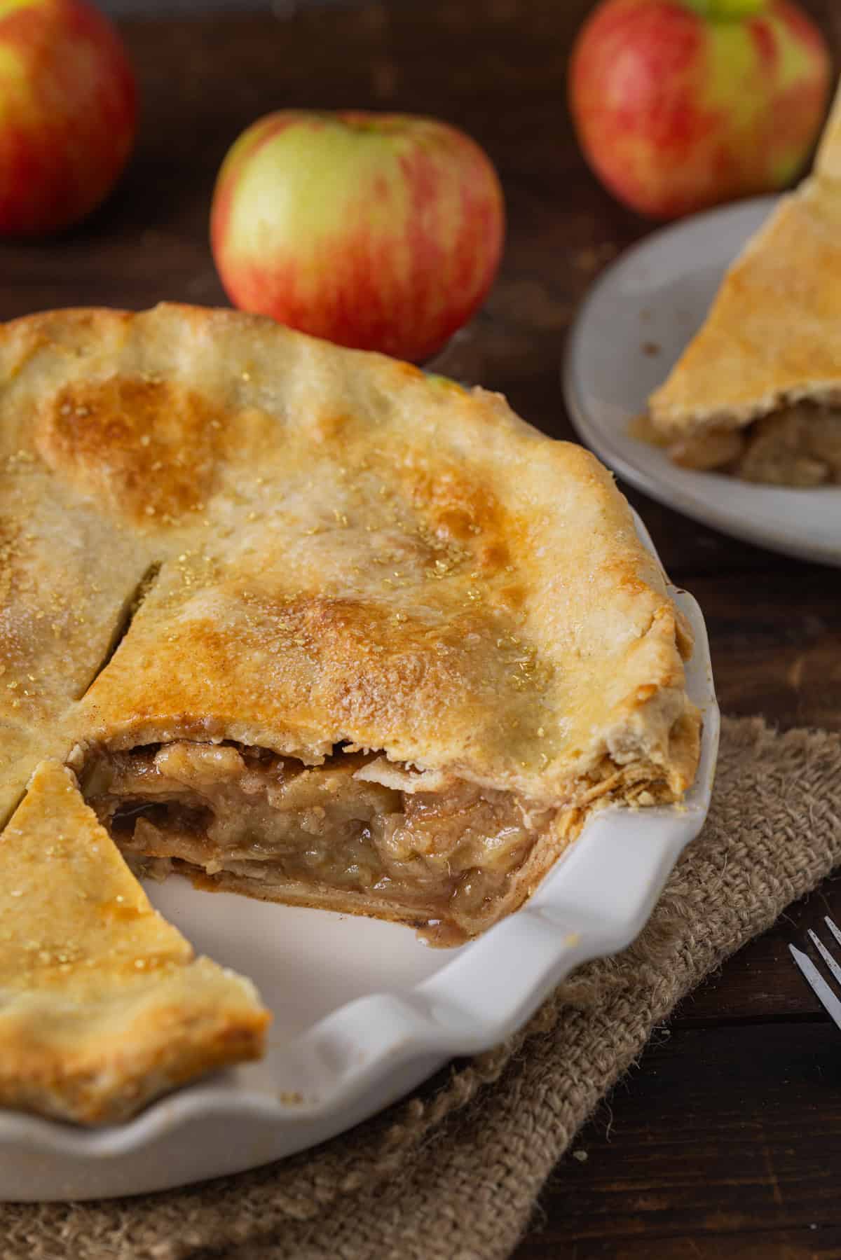 A close-up of an apple pie recipe  with a slice cut out, showing the golden, flaky crust and soft apple filling inside, with whole apples in the background.