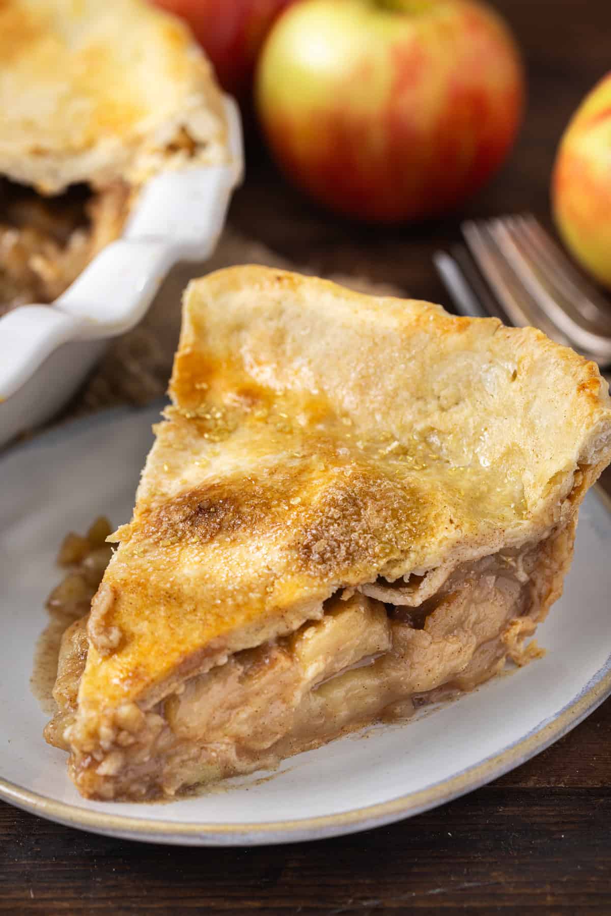 A single slice of apple pie with a golden, flaky crust, served on a white plate, with the rest of the pie and fresh apples in the background.