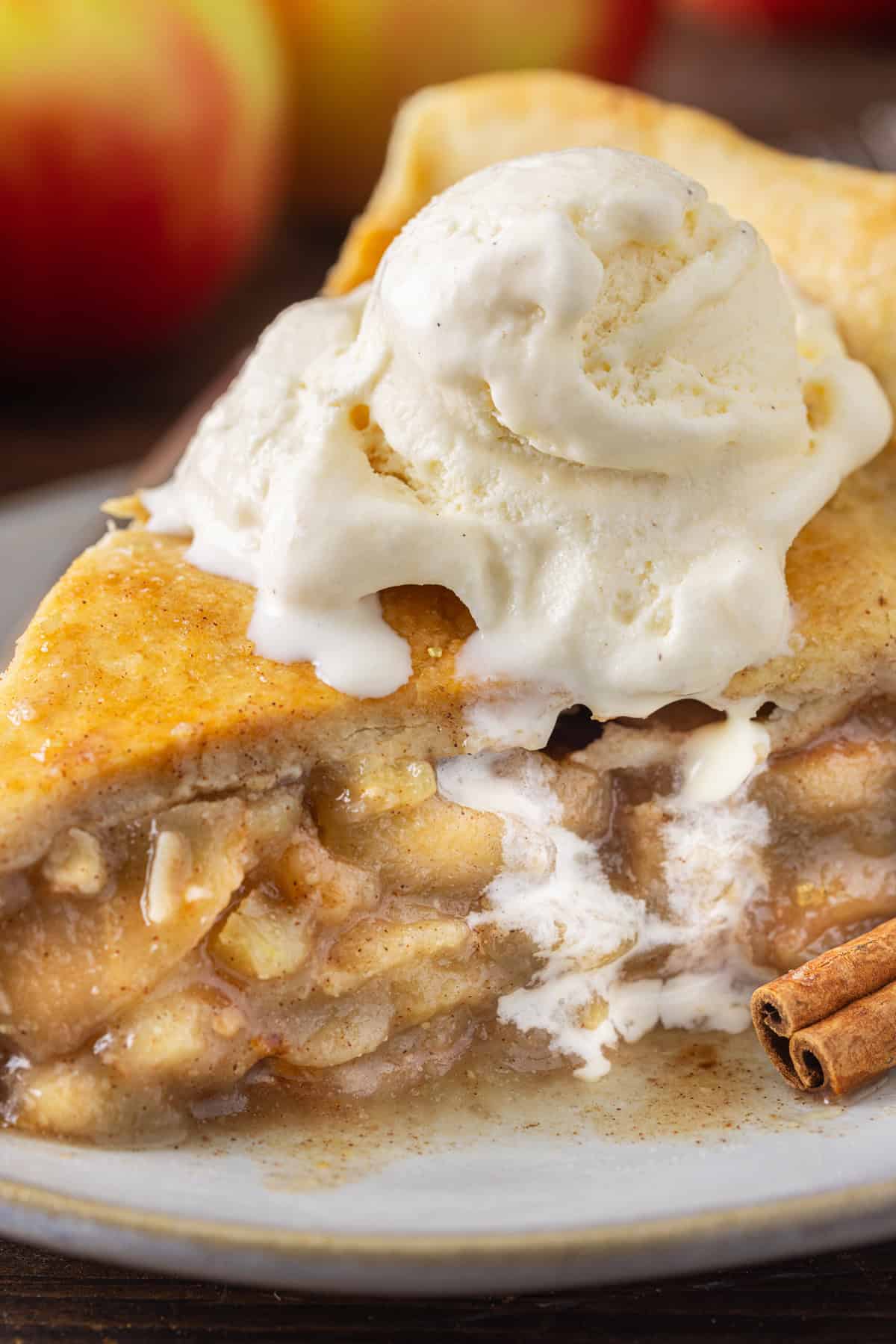 A close-up of the apple pie recipe  slice with a scoop of vanilla ice cream on top, showing the gooey, cinnamon-flavored apple filling and flaky crust, with cinnamon sticks nearby.