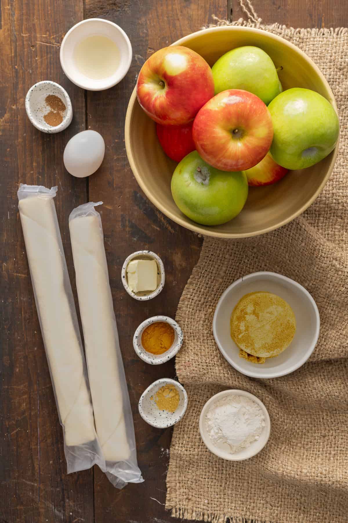 Overhead view of the ingredients for making an apple pie recipe, including whole apples, pie crust, butter, egg, sugar, flour, and cinnamon, all arranged on a rustic wooden surface with burlap.
